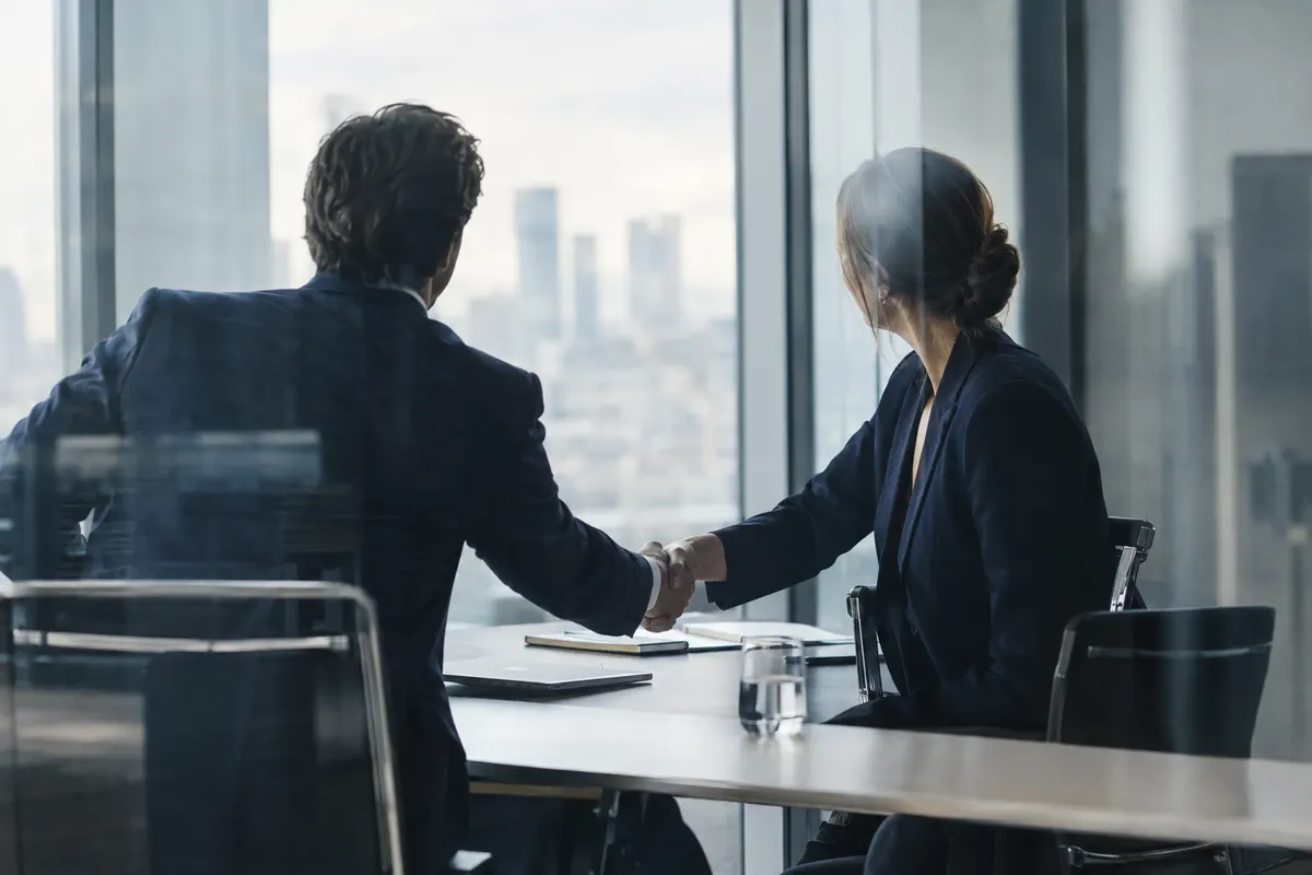 Two consultants shaking hands across a meeting table in a glass-walled conference room